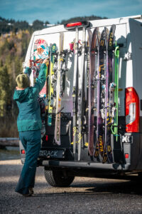 Outdoor enthusiast attaches bikes and skis to PUSH components' Pushrack on a camper van against a mountain backdrop.