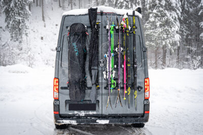 Campervan with PUSHrack ski and snowboard rack, on which several pairs of skis and a snowboard are securely attached to the rear, both with and without bags, in a snowy winter landscape.