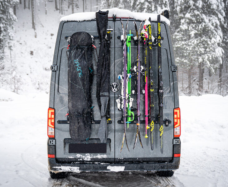 Campervan with PUSHrack ski and snowboard rack, on which several pairs of skis and a snowboard are securely attached to the rear, both with and without bags, in a snowy winter landscape.