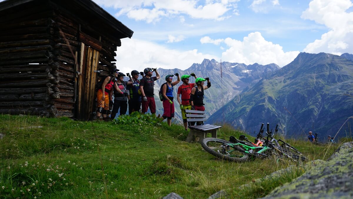 Eine Gruppe Mountainbiker schaut in die Ferne. Im Hintergrund Berge und eine Hütte. Im Vordergrund liegen Mountainbikes.