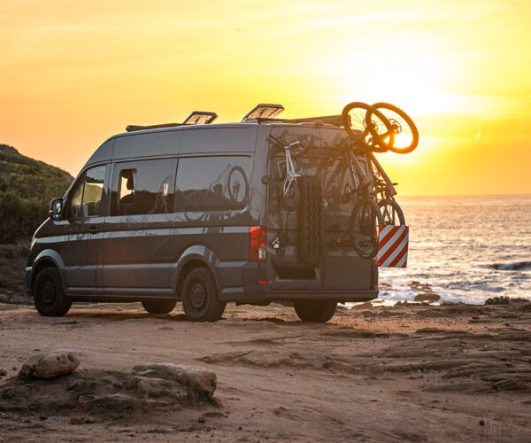 Campervan am Meer bei Sonnenuntergang mit PUSHrack Fahrradträger, zwei Mountainbikes, einem Kinderfahrrad und montierten Auffahrkeilen