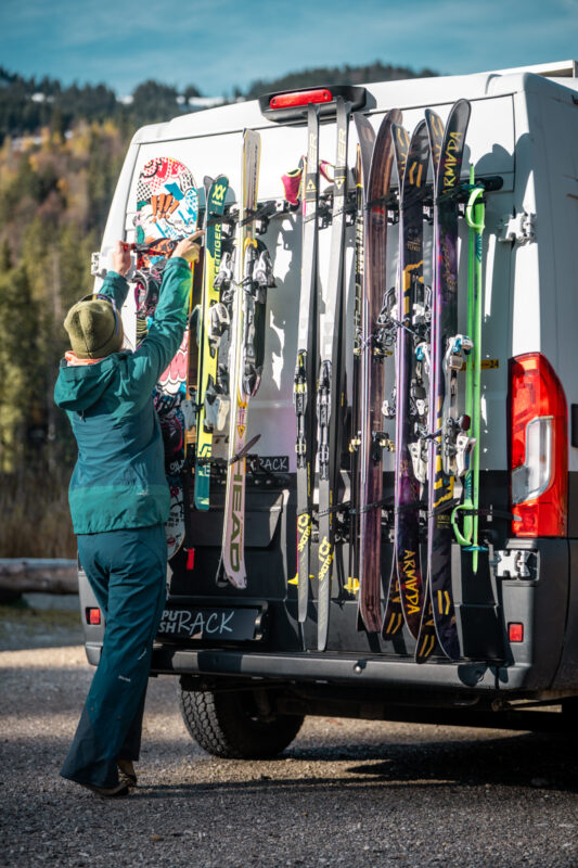 Frau montiert den PUSHrack Snowboardträger am Camper, daneben fünf Paar Ski und zwei Paar Langlaufski am Skiträger