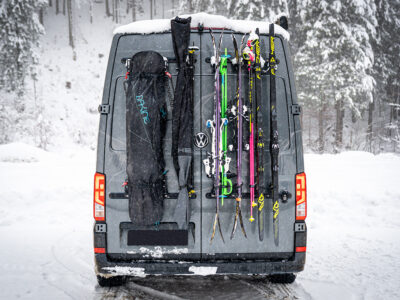 Campervan mit PUSHrack Ski- und Snowboardträger, auf dem mehrere Paar Ski und ein Snowboard sowohl mit als auch ohne Tasche sicher am Heck befestigt sind, in verschneiter Winterlandschaft
