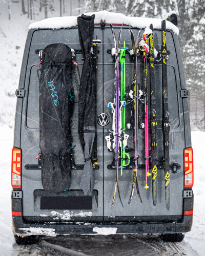 Campervan mit PUSHrack Ski- und Snowboardträger, auf dem mehrere Paar Ski und ein Snowboard sowohl mit als auch ohne Tasche sicher am Heck befestigt sind, in verschneiter Winterlandschaft