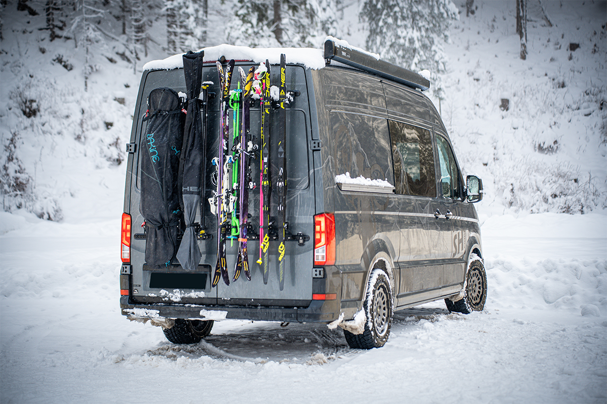Campervan mit PUSHrack Ski- und Snowboardträger, auf dem mehrere Paar Ski und ein Snowboard sowohl mit als auch ohne Tasche sicher am Heck befestigt sind in verschneiter Winterlandschaft