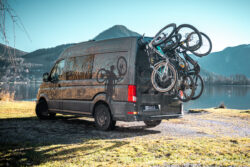 Campervan mit PUSHrack Fahrradträger am Heck, auf dem drei Mountainbikes und ein Kinderfahrrad sicher befestigt sind, geparkt am See vor Berglandschaft