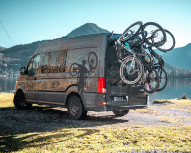 Campervan mit PUSHrack Fahrradträger am Heck, auf dem drei Mountainbikes und ein Kinderfahrrad sicher befestigt sind, geparkt am See vor Berglandschaft
