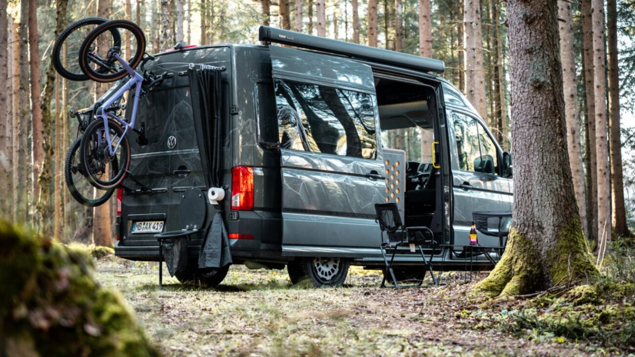 Zu sehen ist ein Camper im Wald, geladen hat er zwei Fahrräder und ein Pupsrack