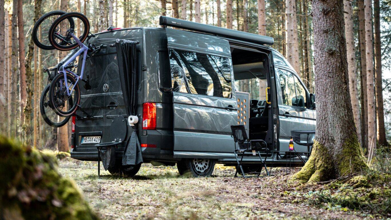 Zu sehen ist ein Camper im Wald, geladen hat er zwei Fahrräder und ein Pupsrack