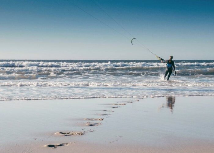 Im Vordergrund sieht man die Fußabdrücke im Sand einer Frau die mit ihrem Kite im Wasser steht